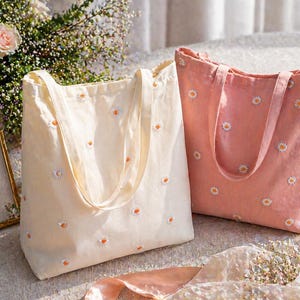 May include: Two tote bags, one cream and one coral, are displayed. Both bags are embroidered with small white and orange daisy flowers. The cream bag is on the left, and the coral bag is on the right. The bags have long handles and are made of a soft fabric.