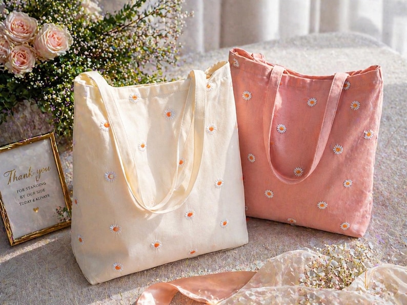 May include: Two tote bags, one cream and one coral, are displayed. Both bags are embroidered with small white and orange daisy flowers. The cream bag is on the left, and the coral bag is on the right. The bags have long handles and are made of a soft fabric.