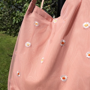 May include: A light pink tote bag with a daisy pattern. The bag is made of a sheer fabric and features embroidered white daisies with orange centers. The bag has a simple strap and is hanging outdoors.