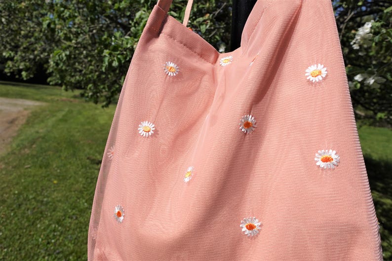 May include: A light pink tote bag with a daisy pattern. The bag is made of a sheer fabric and features embroidered white daisies with orange centers. The bag has a simple strap and is hanging outdoors.