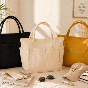 May include: Three canvas tote bags in black, cream, and mustard yellow. Each bag has two exterior pockets and sturdy handles. The mustard bag has the name "Ariana" embroidered in green. A beige baseball cap with "Good day" text is also visible.