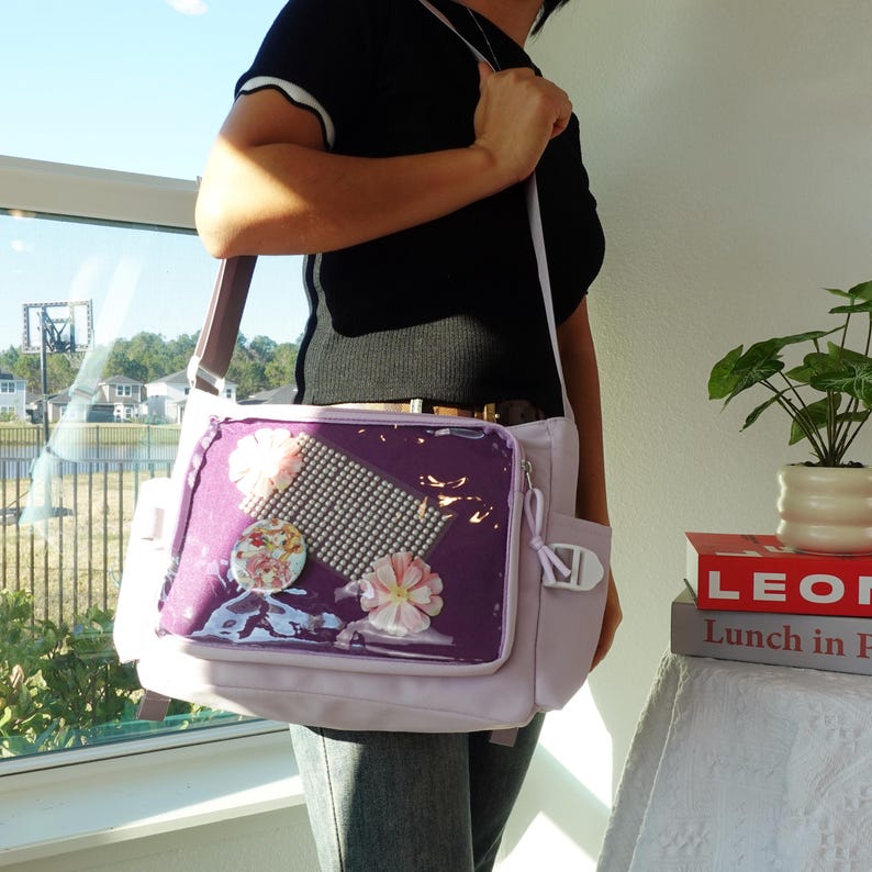 Pu&ograve; includere: Una borsa a tracolla lilla con una tasca frontale trasparente che mostra decorazioni floreali e una griglia di perle. La borsa ha una tracolla marrone e una tasca laterale. La borsa &egrave; portata a spalla.