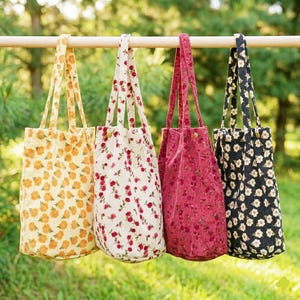 May include: Four corduroy tote bags with floral patterns hang from a wooden rod. The bags are in shades of yellow, white, red, and black. Each bag has a matching handle and a button closure. The bags are ideal for carrying groceries or personal items.