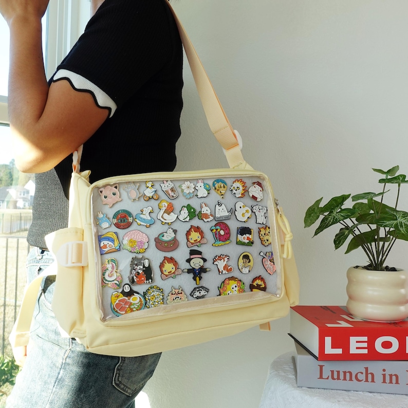 Pu&ograve; includere: Una borsa a tracolla color crema con un pannello frontale trasparente che mostra numerose spille smaltate colorate. La borsa ha una tracolla e una patta frontale con chiusura a fibbia. Le spille presentano vari personaggi e disegni di cartoni animati.