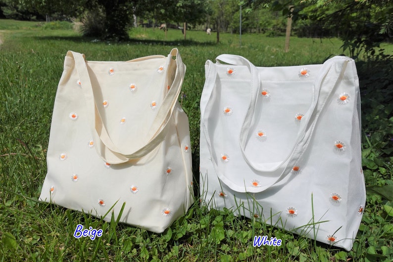 May include: Two canvas tote bags, one beige and one white, are displayed outdoors. Both bags are adorned with embroidered daisy patterns featuring white petals and orange centers. The bags have long straps for shoulder carry.