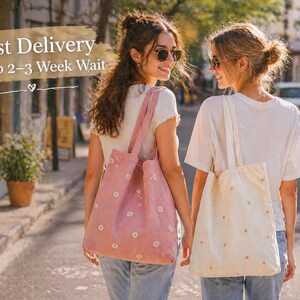 May include: Two women walking down a street, each carrying a tote bag. One bag is pink with white daisy patterns, the other is cream-colored with orange daisy patterns. The text "Fast Delivery Skip 2-3 Week Wait" is visible in the image.