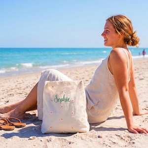 May include: A canvas tote bag with the name "Sophie" in green script and small daisy accents. The bag is on a sandy beach with brown sandals nearby. The scene is set on a sunny day with a clear blue sky and ocean.