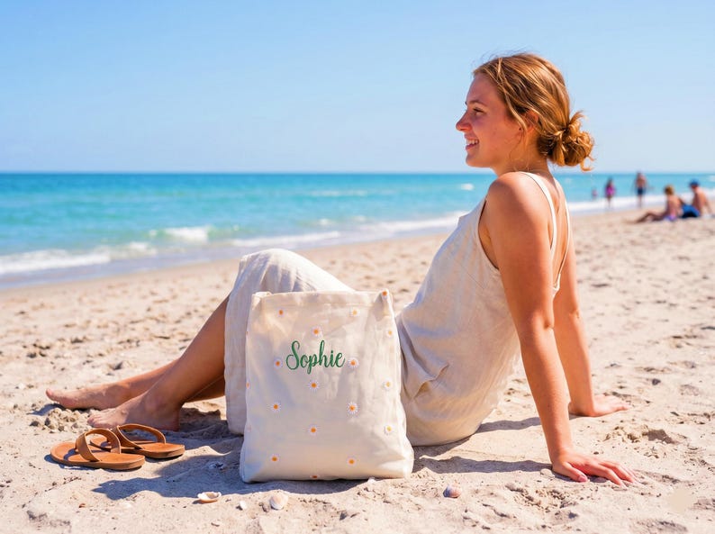 May include: A canvas tote bag with the name "Sophie" in green script and small daisy accents. The bag is on a sandy beach with brown sandals nearby. The scene is set on a sunny day with a clear blue sky and ocean.
