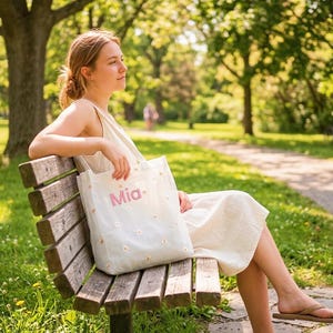 May include: A white tote bag with the name "Mia" in pink script and small daisy flower accents. The bag is held by a person sitting on a wooden park bench. The background features a park setting with trees and a walking path.