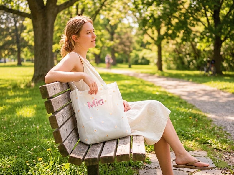 May include: A white tote bag with the name "Mia" in pink script and small daisy flower accents. The bag is held by a person sitting on a wooden park bench. The background features a park setting with trees and a walking path.