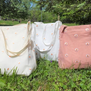 May include: Three canvas tote bags in cream, white, and pink, each adorned with a daisy pattern. The bags have long straps and are displayed on a grassy surface, suggesting an outdoor setting.