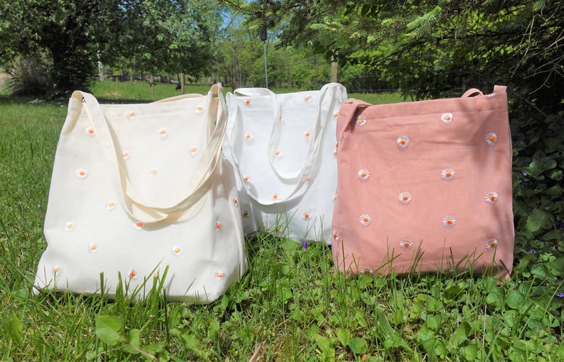 May include: Three canvas tote bags in cream, white, and pink, each adorned with a daisy pattern. The bags have long straps and are displayed on a grassy surface, suggesting an outdoor setting.