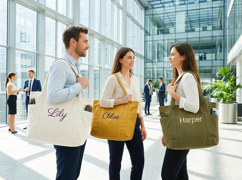 May include: Three people in a modern office setting each carry a personalized tote bag. The bags are in cream, mustard yellow, and olive green, with the names "Lily", "Chloe", and "Harper" embroidered on them. The bags have pockets and are carried over the shoulder.