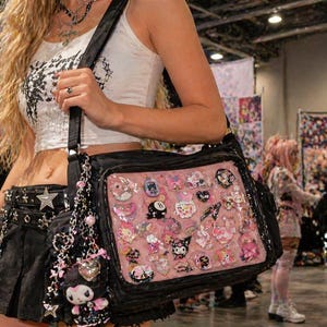 Pu&ograve; includere: Borsa a tracolla nera con un pannello frontale rosa pieno di varie spille e ciondoli decorativi. La borsa ha una tracolla nera e un ciondolo pendente. La borsa &egrave; portata da una persona che indossa un top corto bianco e una gonna nera.