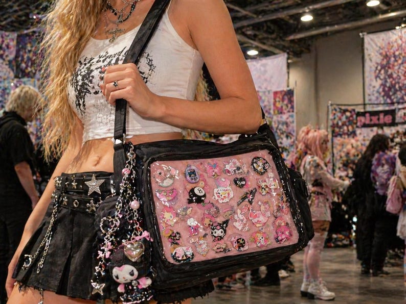 Pu&ograve; includere: Borsa a tracolla nera con un pannello frontale rosa pieno di varie spille e ciondoli decorativi. La borsa ha una tracolla nera e un ciondolo pendente. La borsa &egrave; portata da una persona che indossa un top corto bianco e una gonna nera.