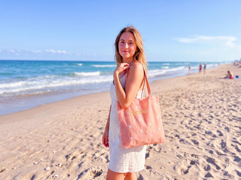 May include: A coral-colored tote bag with white daisy embroidery and the word "bonnie" in gold lettering. The bag has brown straps and is held by a person on a sandy beach with the ocean in the background.