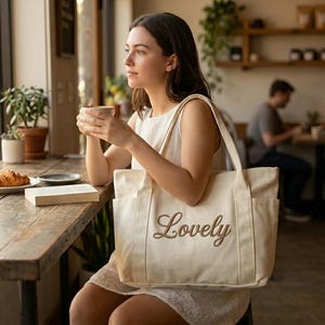 May include: A canvas tote bag with the word "Lovely" embroidered in a cursive font. The bag is cream-colored and has two exterior pockets. The image is taken in a cafe setting, with a woman holding the bag.