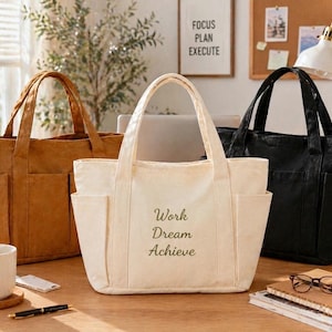Może przedstawiać: Trzy torby typu tote w różnych kolorach: brązowym, kremowym i czarnym. Kremowa torba ma nadruk "Work Dream Achieve". Każda torba ma dwie zewnętrzne kieszenie i solidne uchwyty. Torby leżą na drewnianej powierzchni.