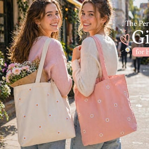 May include: Two women holding tote bags with daisy embroidery. One bag is cream-colored, the other is pink. The bags have long straps and are filled with flowers. The text "The Perfect Gift for Her" is visible.