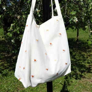May include: A white tote bag with a daisy pattern. The bag has two long straps and is made of a soft fabric. The daisies are white with yellow centers and are scattered across the bag.
