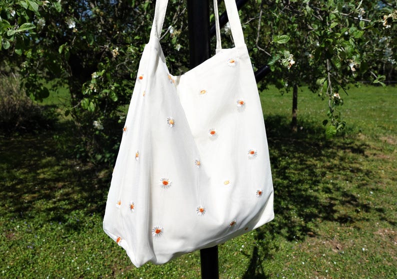 May include: A white tote bag with a daisy pattern. The bag has two long straps and is made of a soft fabric. The daisies are white with yellow centers and are scattered across the bag.
