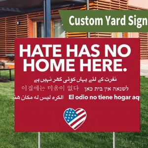 May include: A red yard sign with the message "HATE HAS NO HOME HERE" in white text. The sign includes text in multiple languages and an American flag heart graphic. The sign is displayed in a grassy garden.