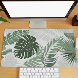 Puede incluir: Una alfombrilla de escritorio blanca con un patrón de hojas tropicales verdes. La alfombrilla está sobre un escritorio de madera con un monitor de ordenador, un teclado y un ratón.