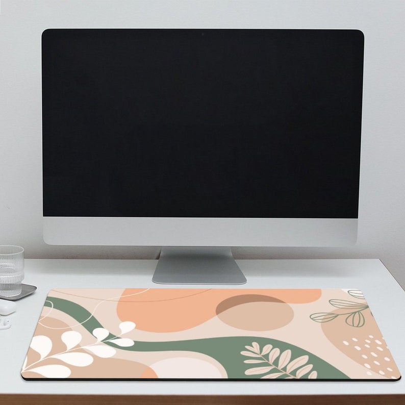 May include: A beige desk mat with an abstract design featuring green, peach, and brown shapes and white leaves. The mat is on a white desk with a computer monitor on top.