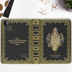 Peut inclure: Une coque de téléphone noire avec un motif doré. La coque présente un design de livre avec une bordure dorée ornée et une citation qui dit "FEAR CAN HOLD YOU PRISONER HOPE CAN SET YOU FREE". Le dos de la coque présente un blason doré et le texte "SALVATION LIES WITHIN".