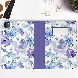 Puede incluir: Una funda de teléfono blanca con un patrón floral morado. La funda tiene una solapa que cubre la pantalla y una ranura para una tarjeta.