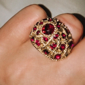 Puede incluir: Un anillo de tono dorado con una superficie de metal texturizado y trenzado, incrustado con numerosas piedras pequeñas, redondas y de color rojo intenso facetadas. El diseño tiene forma de cúpula, con una piedra central grande y otras pequeñas.