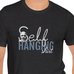 Pode incluir: Uma camiseta cinza escura com um design de corda branca que diz "Self Hanging shirt". O rosto de um homem está dentro do design da corda.
