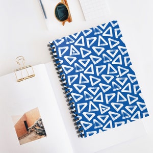 Puede incluir: Una libreta espiral azul con un patrón geométrico blanco de triángulos. La libreta está sobre una superficie blanca con un clip de carpeta dorado y unas gafas de sol.