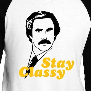 May include: A white and black baseball style t-shirt with a black and white illustration of a man with a mustache. The text "Stay Classy" is printed in yellow on the shirt.