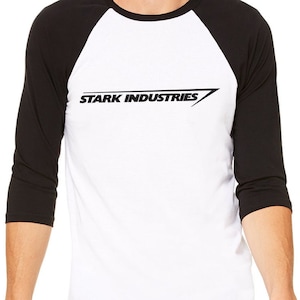 Puede incluir: Una camiseta de b&eacute;isbol blanca y negra con un logotipo negro que dice "STARK INDUSTRIES" en una fuente inclinada.