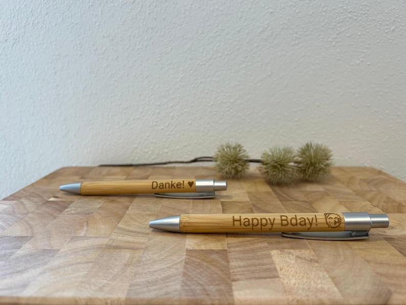 Puede incluir: Dos bol&iacute;grafos de bamb&uacute; con detalles plateados. El bol&iacute;grafo superior tiene el texto "Danke!" con un s&iacute;mbolo de coraz&oacute;n. El bol&iacute;grafo inferior tiene el texto "Happy Bday!" con un peque&ntilde;o s&iacute;mbolo de huella de pata.