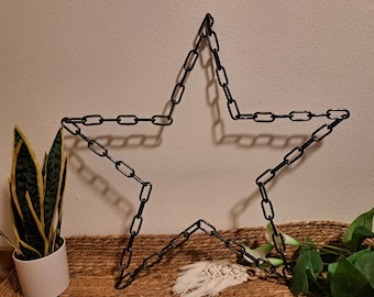 Puede incluir: Una estrella de metal negro hecha de eslabones de cadena. La estrella se muestra contra una pared neutra, con una estera tejida y vegetación en primer plano. La estrella es una pieza decorativa.