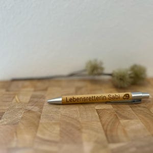 Puede incluir: Un bol&iacute;grafo de madera con una punta plateada y el texto "Lebensretterin Sabi" grabado en &eacute;l. El bol&iacute;grafo est&aacute; sobre una superficie de madera.