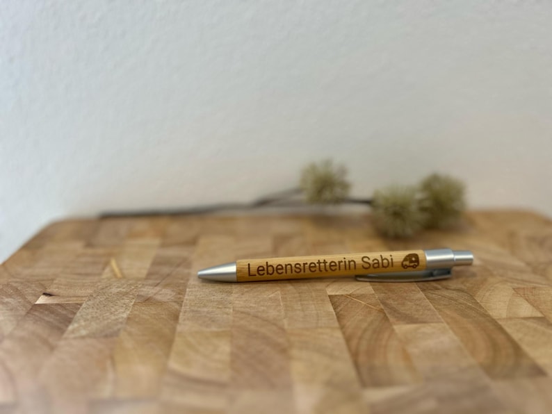 Puede incluir: Un bol&iacute;grafo de madera con una punta plateada y el texto "Lebensretterin Sabi" grabado en &eacute;l. El bol&iacute;grafo est&aacute; sobre una superficie de madera.