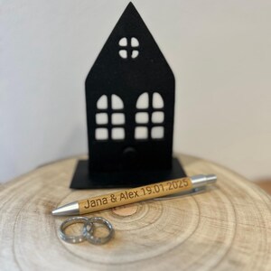 Puede incluir: Un soporte negro con forma de casa con un bol&iacute;grafo de madera que dice "Jana & Alex 19.01.2025" y dos anillos plateados sobre una superficie de madera.