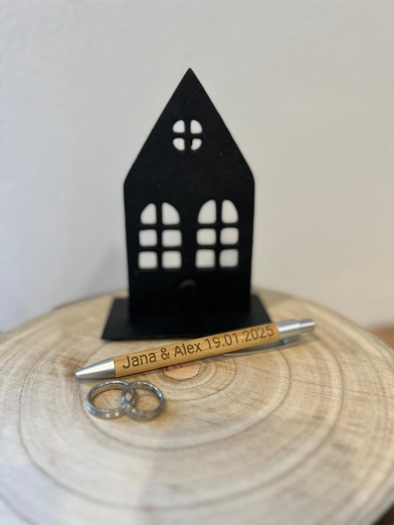 Puede incluir: Un soporte negro con forma de casa con un bol&iacute;grafo de madera que dice "Jana & Alex 19.01.2025" y dos anillos plateados sobre una superficie de madera.