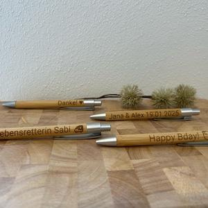 Puede incluir: Cuatro bol&iacute;grafos de madera con detalles plateados y mensajes personalizados. Los mensajes incluyen "Danke!" con un coraz&oacute;n, "Lebensretterin Sabi" con un peque&ntilde;o icono de coche, "Jana & Alex 19.01.2025" y "Happy Bday!" con un peque&ntilde;o icono de perro.