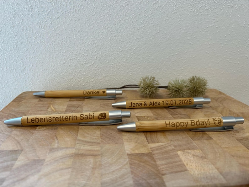 Puede incluir: Cuatro bol&iacute;grafos de madera con detalles plateados y mensajes personalizados. Los mensajes incluyen "Danke!" con un coraz&oacute;n, "Lebensretterin Sabi" con un peque&ntilde;o icono de coche, "Jana & Alex 19.01.2025" y "Happy Bday!" con un peque&ntilde;o icono de perro.