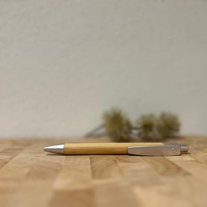 Puede incluir: Un bol&iacute;grafo de madera con un clip plateado y una punta plateada. El bol&iacute;grafo est&aacute; sobre una superficie de madera.