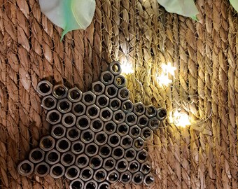Puede incluir: Un adorno en forma de estrella hecho de numerosas tuercas metálicas, sobre un fondo tejido texturizado. La estrella está rodeada de pequeñas luces blancas cálidas y flores artificiales blancas y verdes.