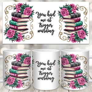 May include: White ceramic mug with a floral design and the text "You had me at trigger warning". The design features a stack of books with pink and green flowers.