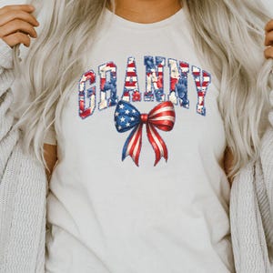 Pode incluir: Camiseta creme com a palavra "GRANNY" em uma fonte patriótica com padrões florais e listrados vermelhos, brancos e azuis. Um laço com listras vermelhas e brancas e estrelas azuis está centralizado abaixo.