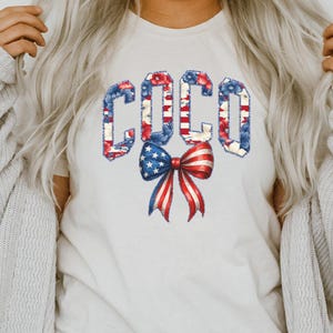 May include: A cream-colored t-shirt featuring the word "COCO" in a patriotic design. The letters are filled with red, white, and blue floral and striped patterns. A bow with red and white stripes and blue stars is below the word.