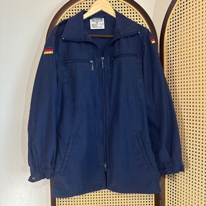 May include: A navy blue jacket with a full-length zip, two chest pockets, and two lower pockets. The jacket has a stand-up collar and German flag patches on the shoulders. A label is visible on the inside of the collar.