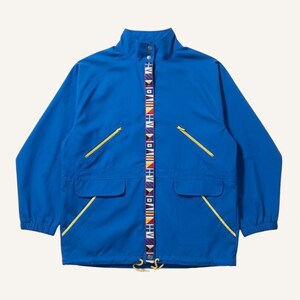May include: A vibrant blue jacket with a stand-up collar and two front pockets. The jacket features yellow zippers and a decorative strip down the center with colorful flag-like patterns. The jacket has long sleeves and a drawstring at the bottom.